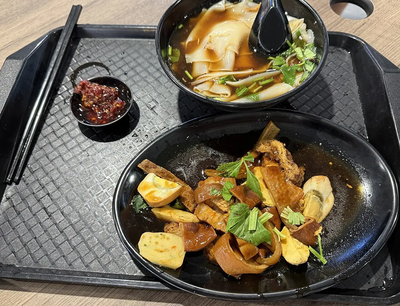 A black tray holds flat rice noodles in broth beside a plate of dark braised food and pale tofu pieces.