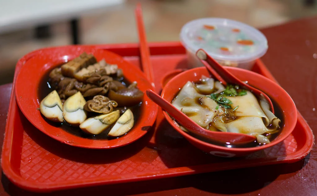 A red tray holds two bowls: one with dark braised pork, egg and tofu, and one with flat rice noodles in soup.