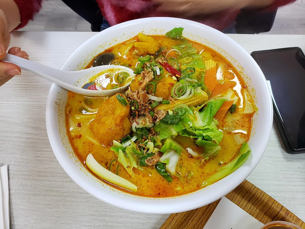 A white bowl of spicy coconut noodle soup with tofu puffs, green vegetables and a spoon lifting broth.