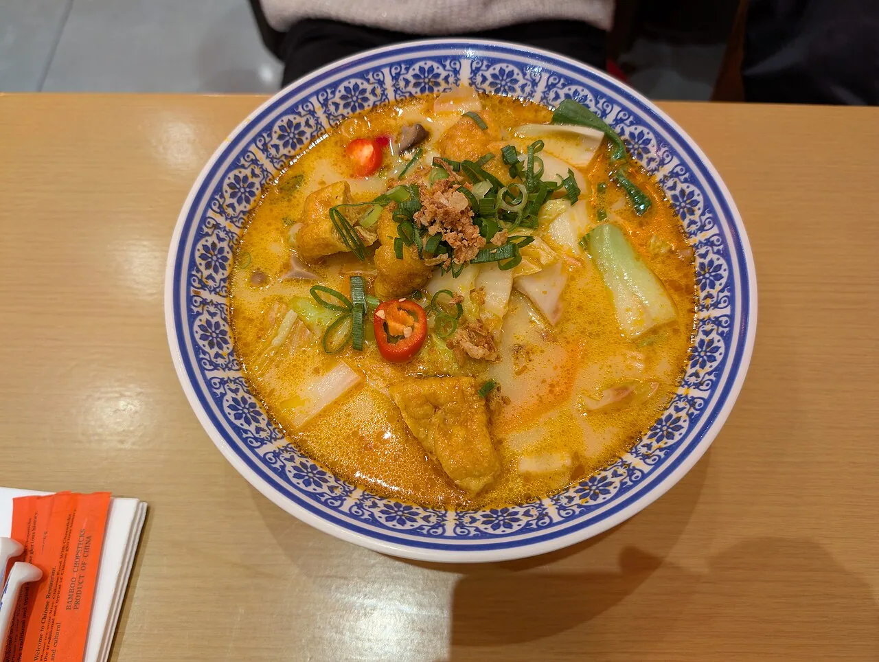 A blue-and-white bowl of orange coconut noodle soup topped with tofu puffs, greens and chilli.