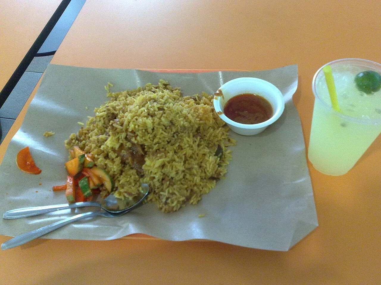 A glass of iced lime juice with a lime wedge beside a plate of mutton biryani.