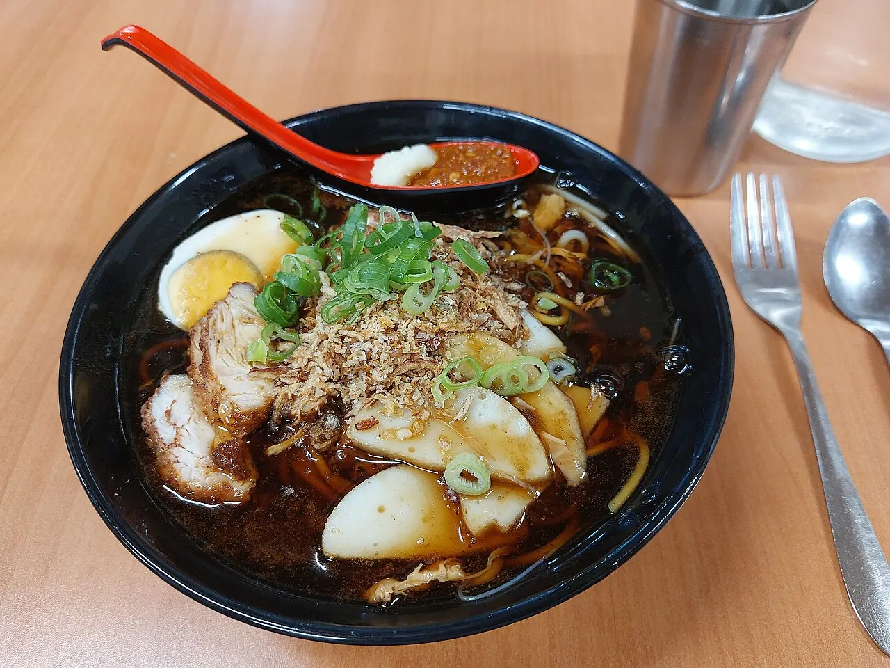 A black bowl of dark noodle soup is topped with sliced egg, fish cake, meat, fried shallots and green onion.