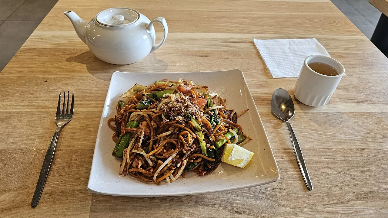 A square plate of dark fried noodles with bean sprouts, green vegetables and a lime wedge on a wooden table.