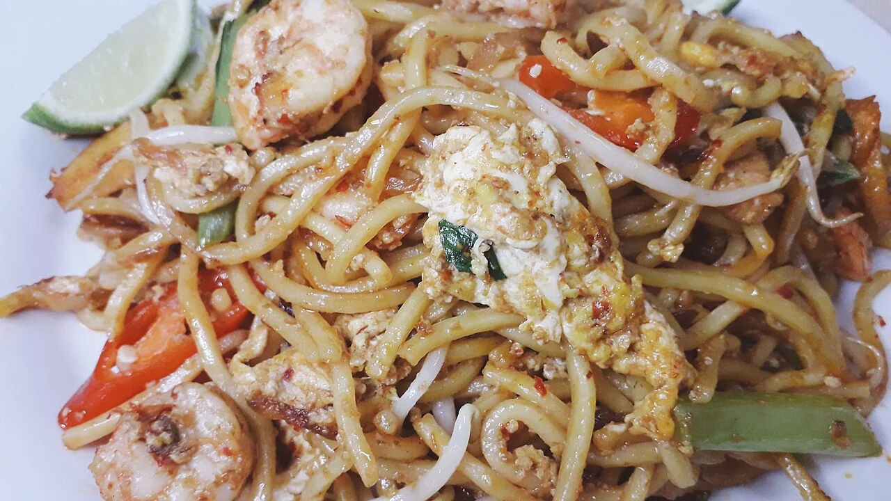 A close-up of yellow fried noodles mixed with egg, prawns, bean sprouts, red pepper and green vegetables on a white plate.