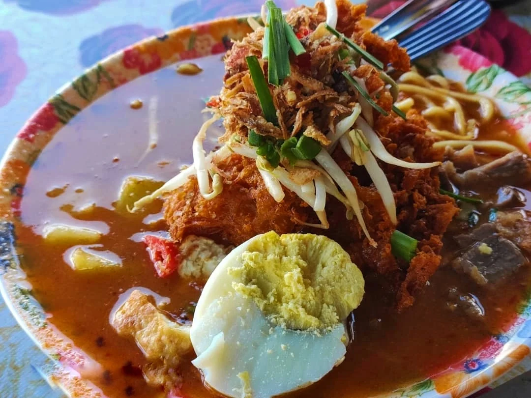A plate of noodles in orange gravy is topped with a halved boiled egg, bean sprouts, fried bits and green onion.
