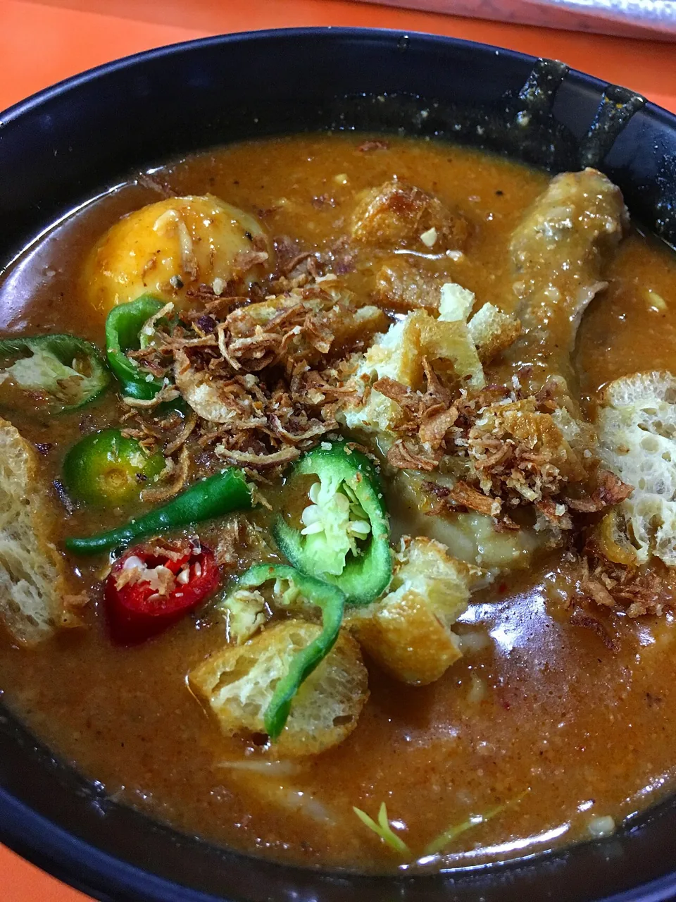 A bowl of yellow noodles in thick brown gravy topped with a boiled egg, fried tofu puffs, green chillies and crispy shallots.