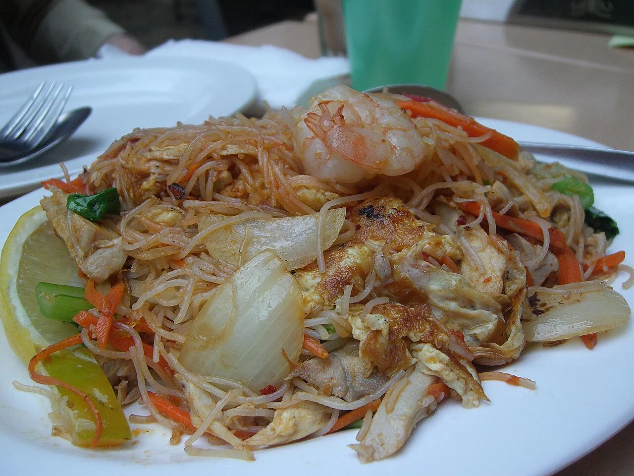 A plate of thin rice noodles with prawns, egg, carrots, green vegetables and a lemon wedge.