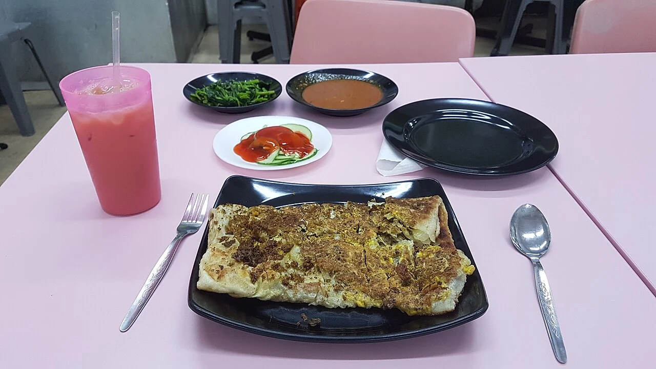 A large rectangular murtabak rests on a black plate beside small dishes of sauce and a pink iced drink.