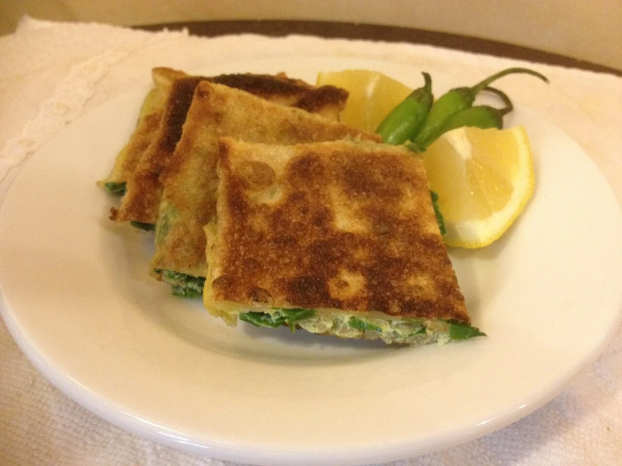Three golden-brown slices of stuffed murtabak on a white plate with lemon and green chillies.