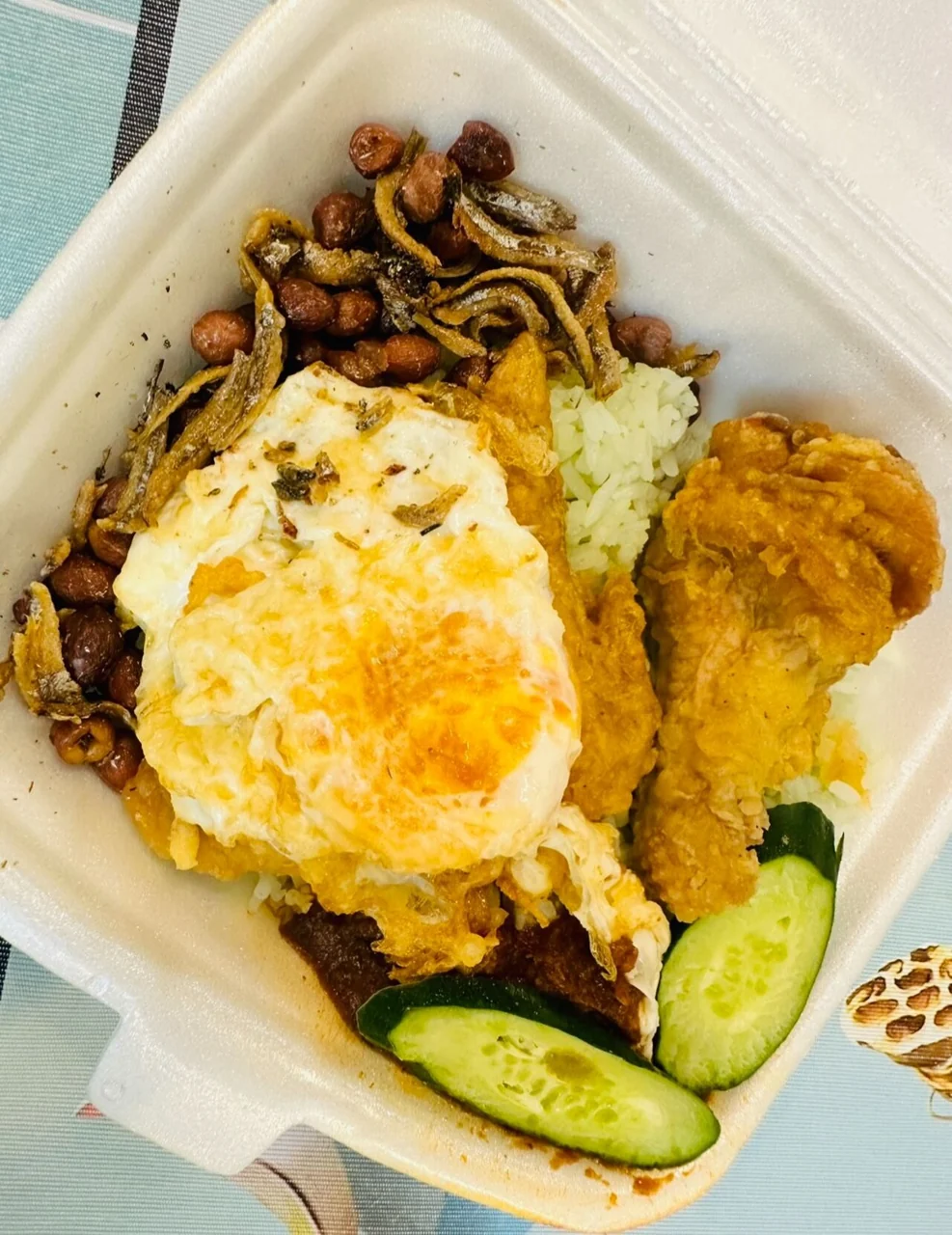 A takeaway box of rice with fried egg, anchovies, peanuts, cucumber slices and a large piece of fried chicken.