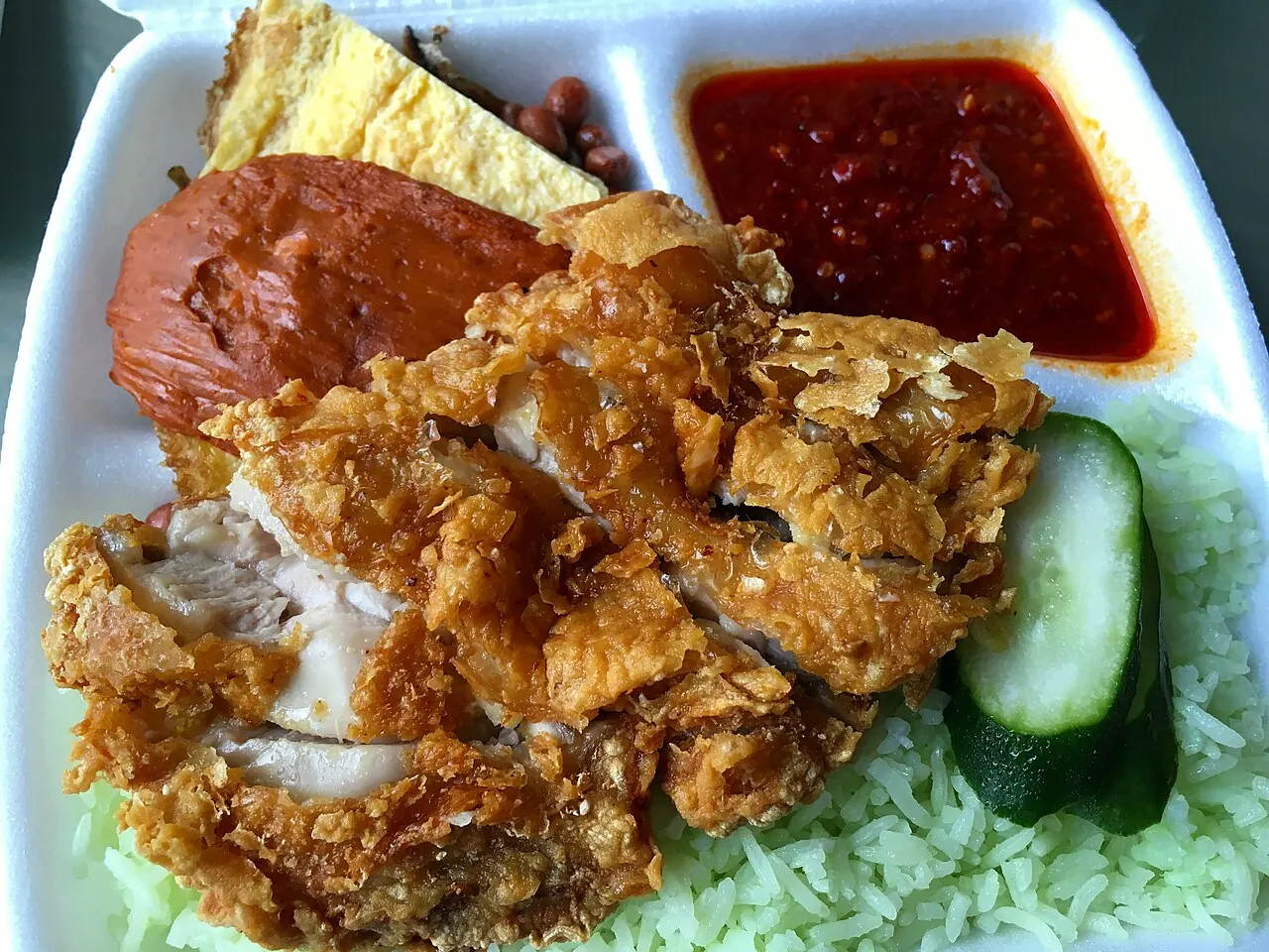 A white box of coconut rice topped with sliced fried chicken, red sambal, egg, peanuts, anchovies and cucumber.