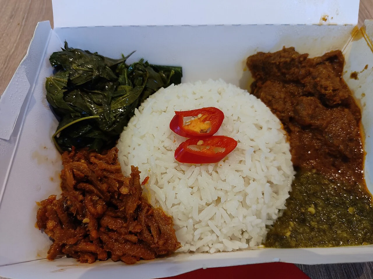 White rice sits between dark green vegetables and several brown and red side dishes in a takeaway box.