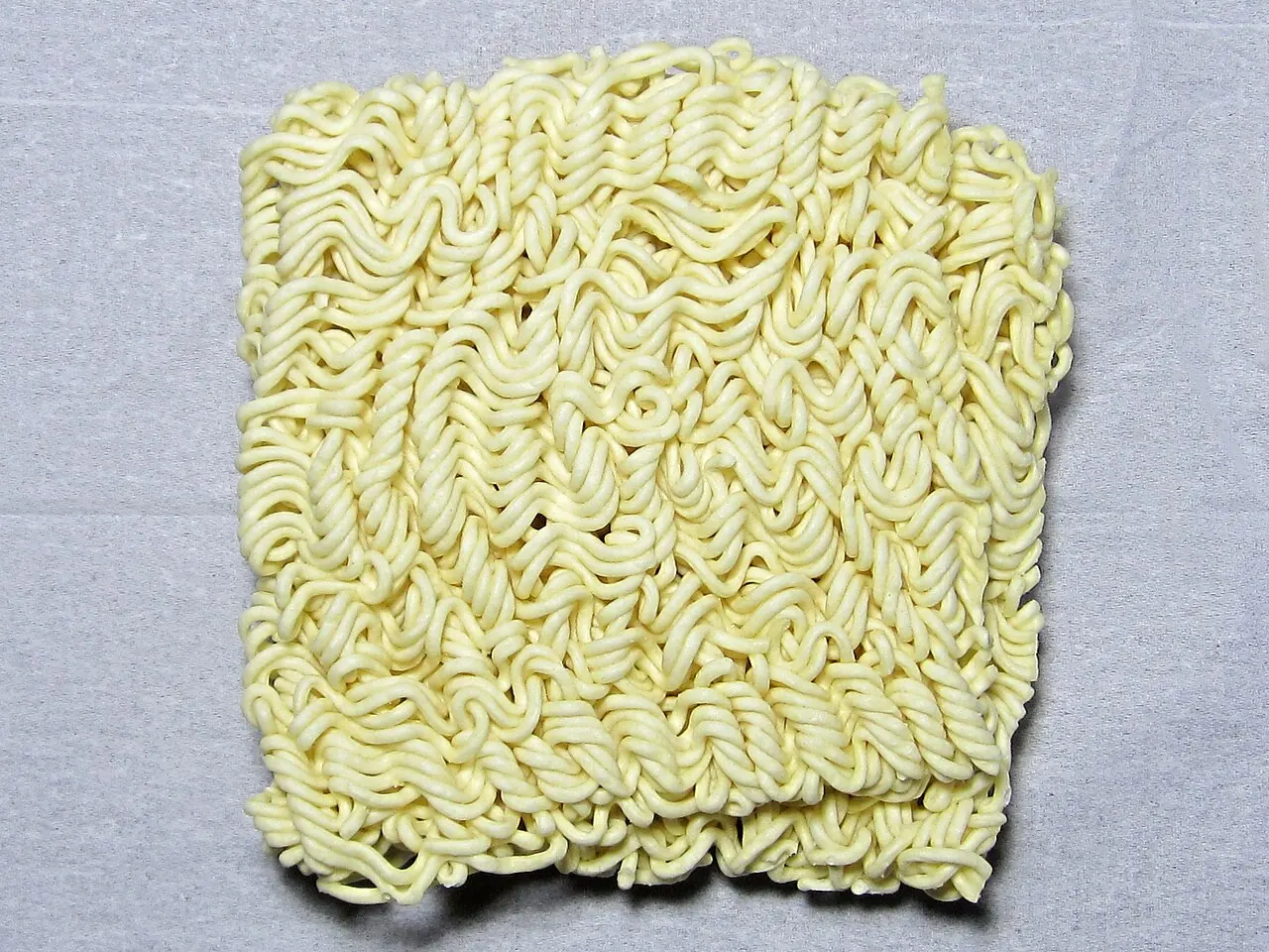A square block of dry wavy instant noodles rests on a light surface.