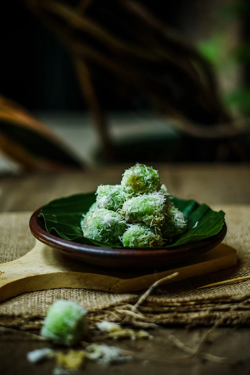 A small pile of green rice balls covered in grated coconut rests on a banana leaf on a dark plate.