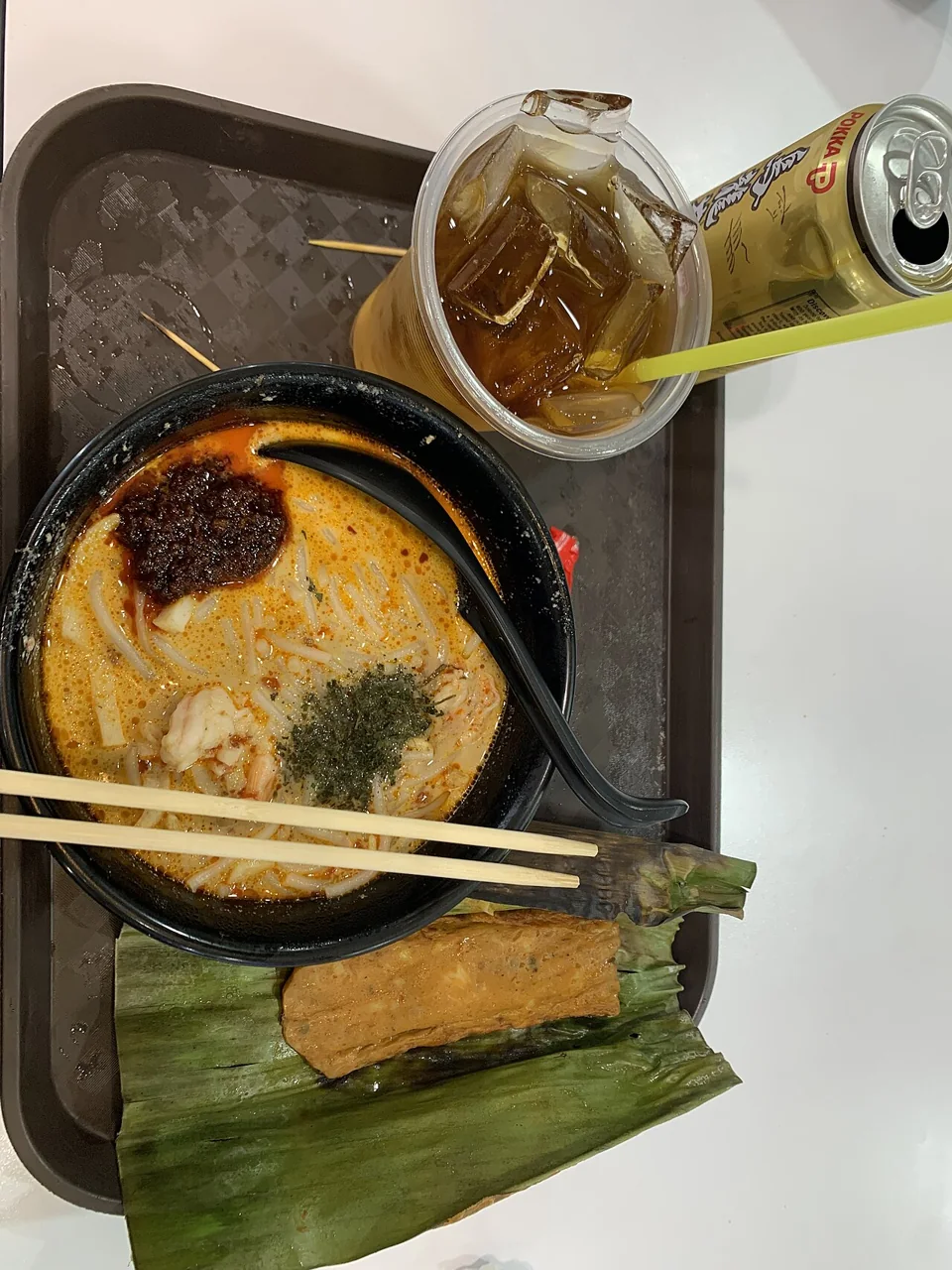 Otah rests on an opened green leaf beside a bowl of laksa and iced tea.