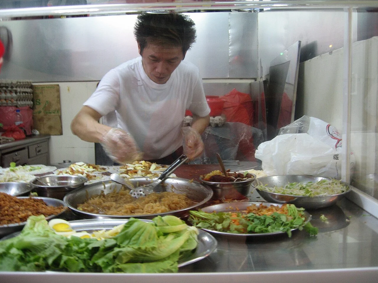 A man in a white shirt prepares popiah at a stall, with lettuce, egg and fillings on the counter.