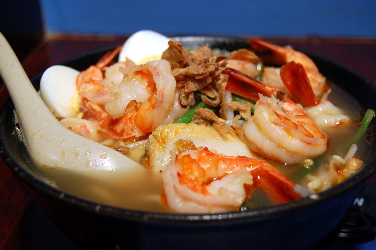 A bowl of prawn noodle soup with large prawns, egg halves, fried shallots and a spoon.