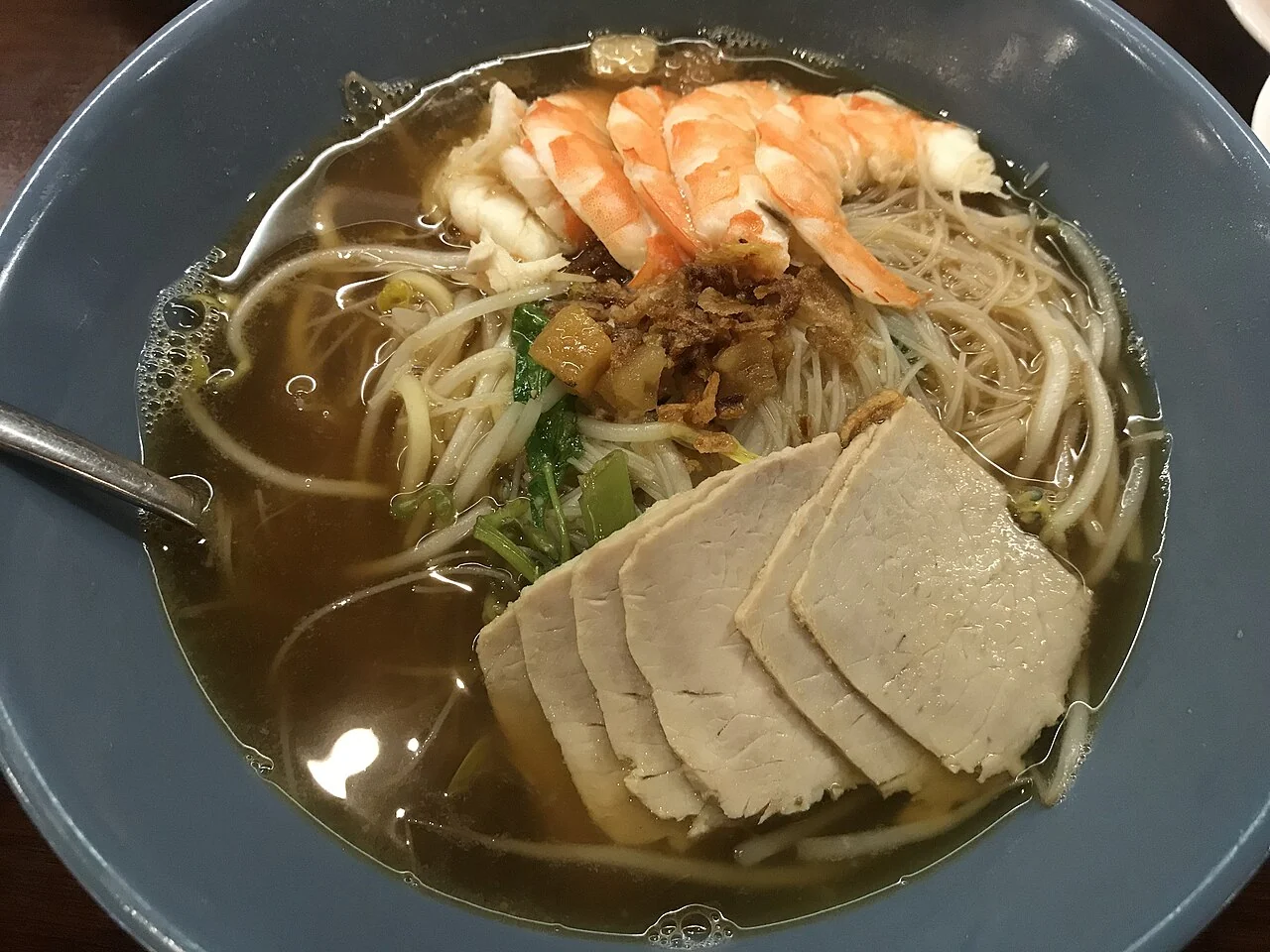 A blue bowl holds noodle soup with orange prawns, sliced pork, bean sprouts and green vegetables in clear broth.