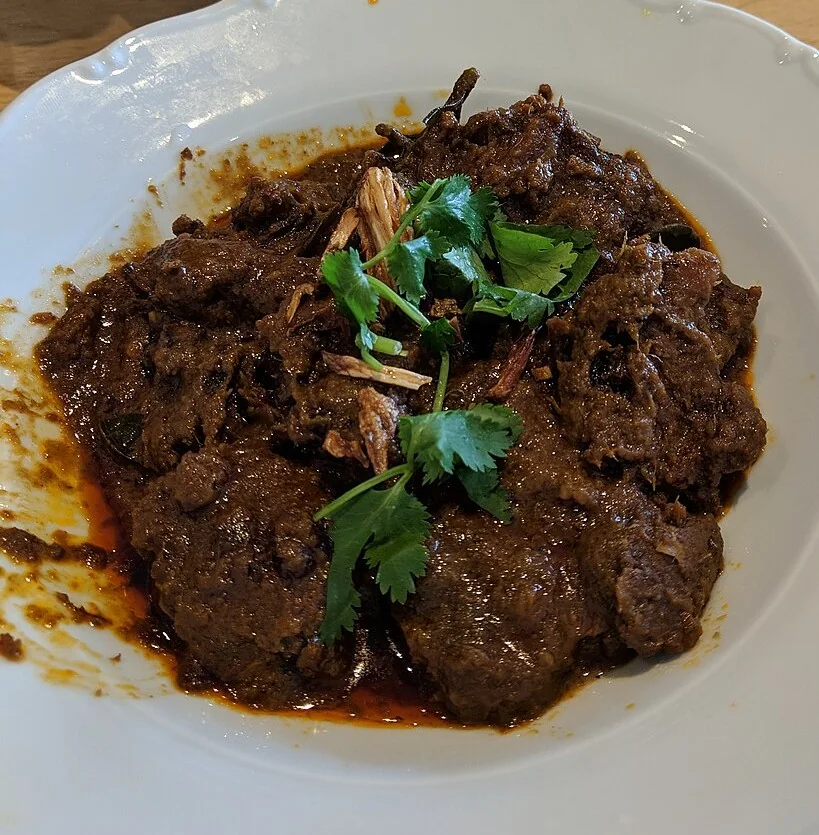 A white plate holds pieces of dark brown beef rendang topped with fresh coriander leaves.