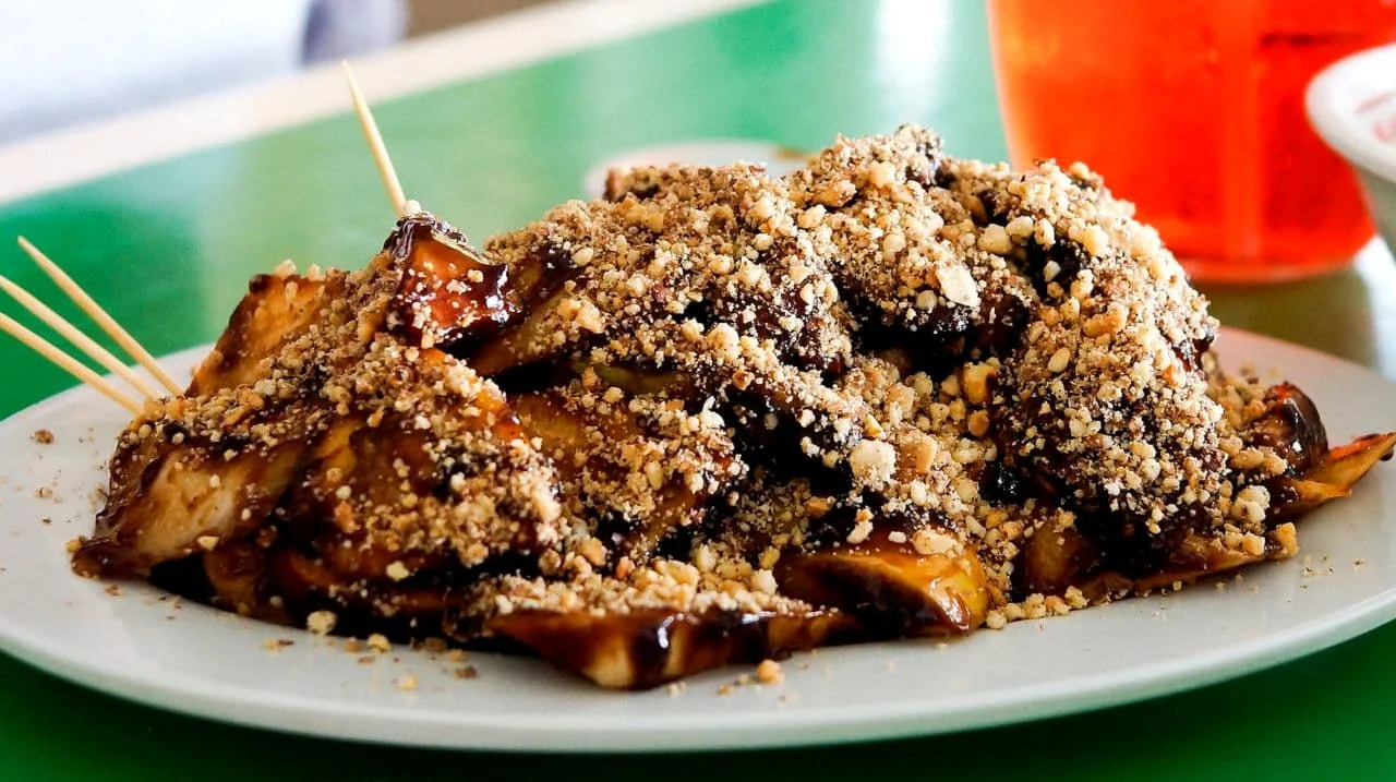 Chunks of fruit covered in dark sauce and crushed peanuts sit on a white plate with wooden skewers.