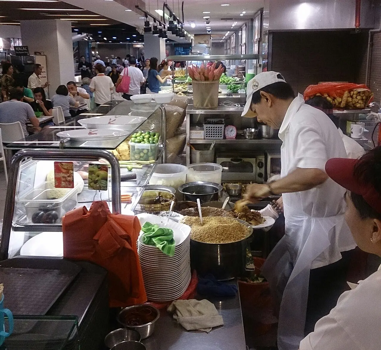 A vendor in a white shirt and cap mixes a plate of rojak beside bowls of peanuts and sauce at a busy food stall.