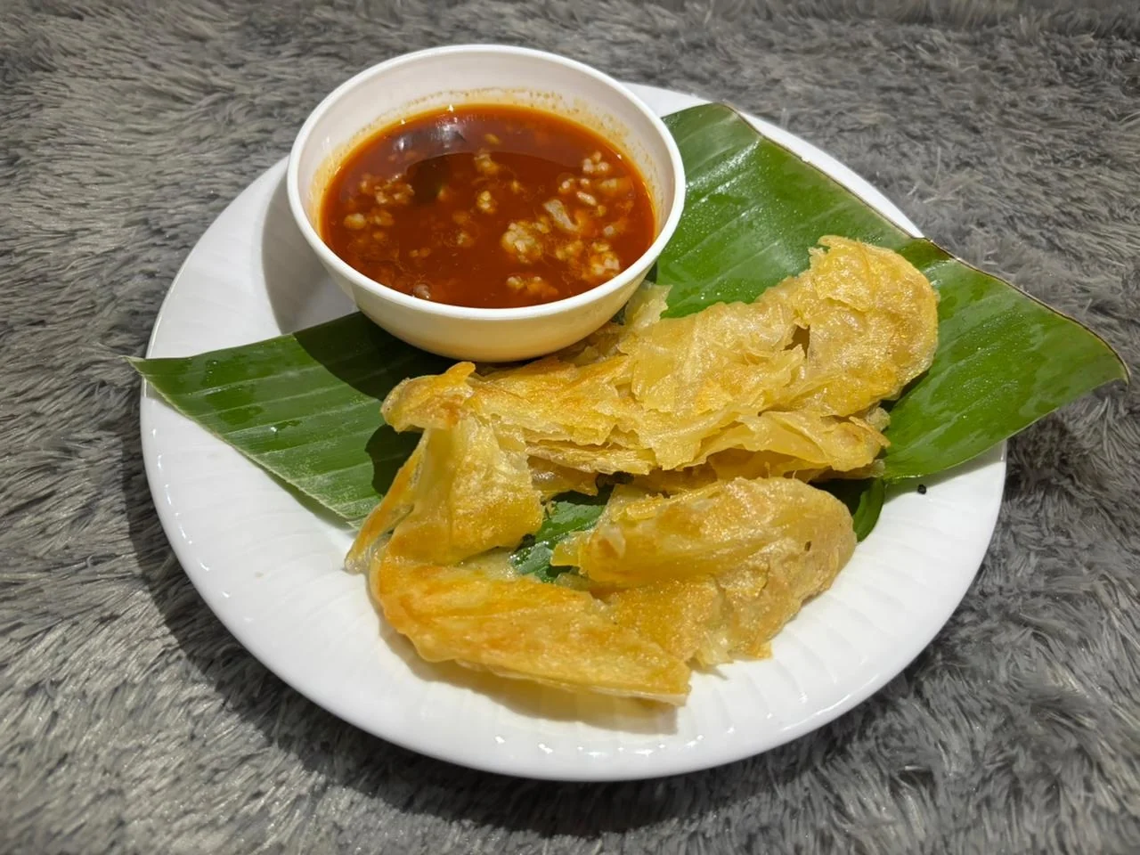 Crispy golden roti prata pieces on a banana leaf, served with a small bowl of red dipping sauce.