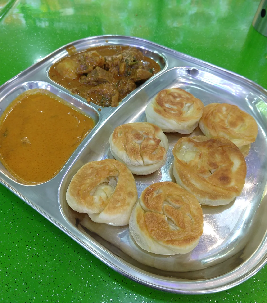 Small round pieces of prata sit on a shiny metal tray beside two portions of curry.