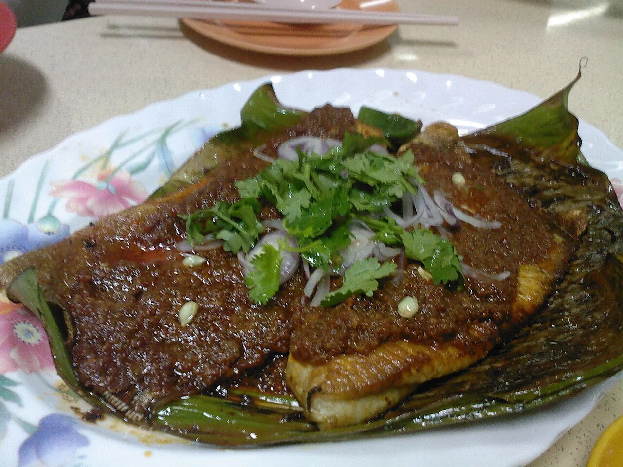 A grilled stingray is served on a green leaf, topped with dark sauce, sliced red onion and fresh coriander leaves.