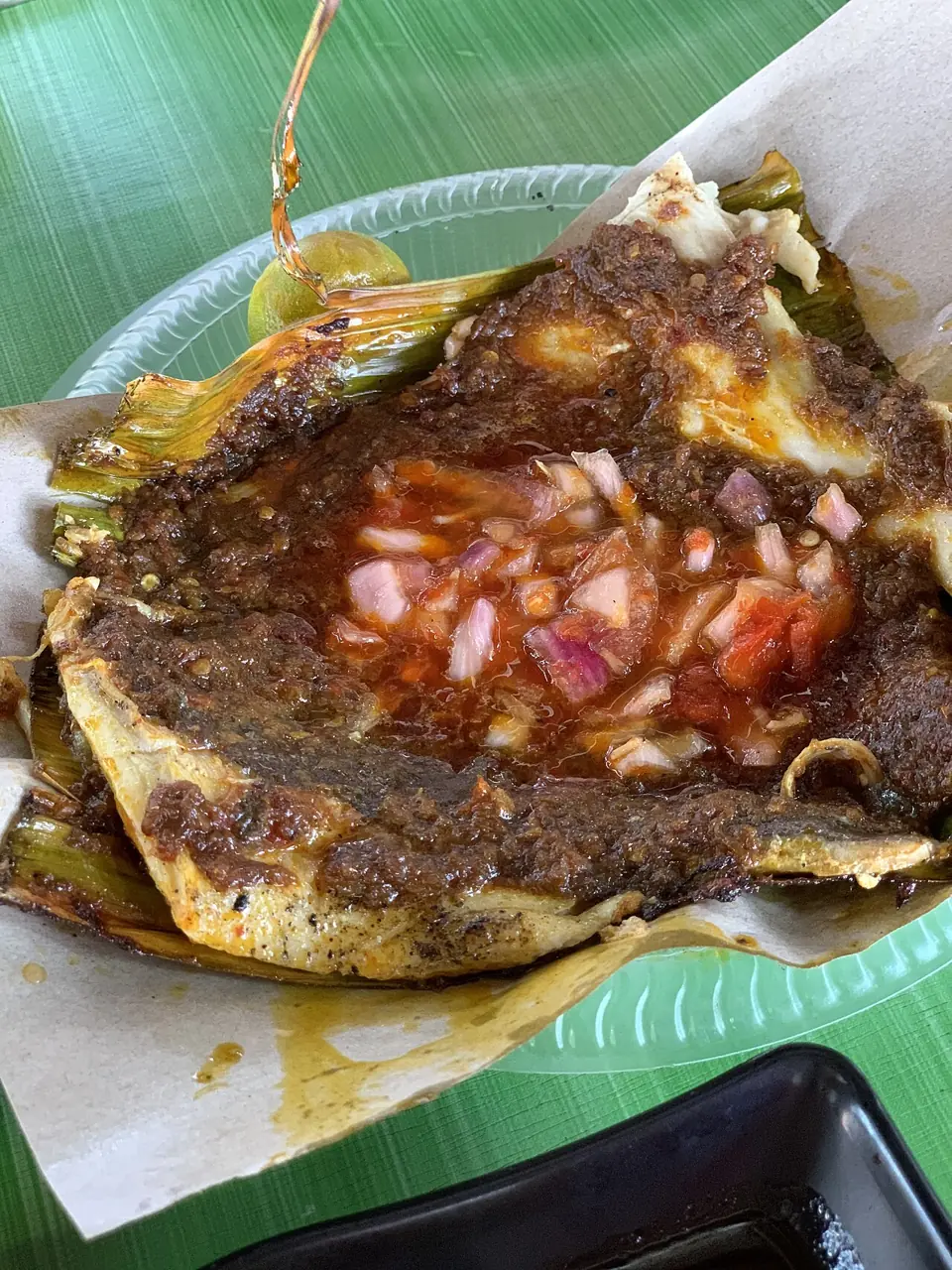 A grilled stingray wing is coated with dark sambal, topped with chopped onion and tomato, and served on banana leaf.