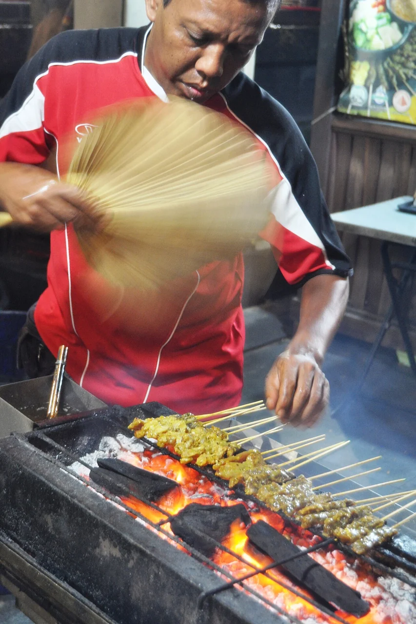 A man in a red shirt fans skewers of satay cooking over a glowing charcoal grill.