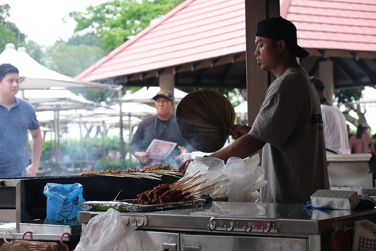A stallholder turns satay skewers over a grill, with other people standing behind the stall.