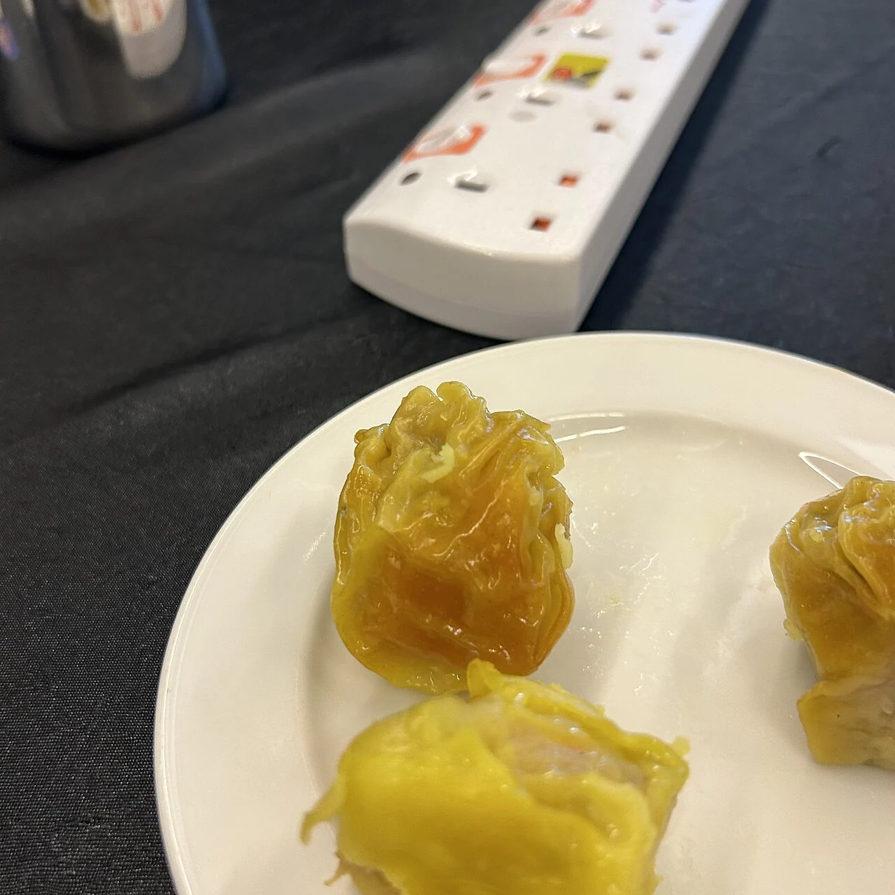 Yellow siew mai dumplings sit on a white plate, with a white power strip behind them on the table.
