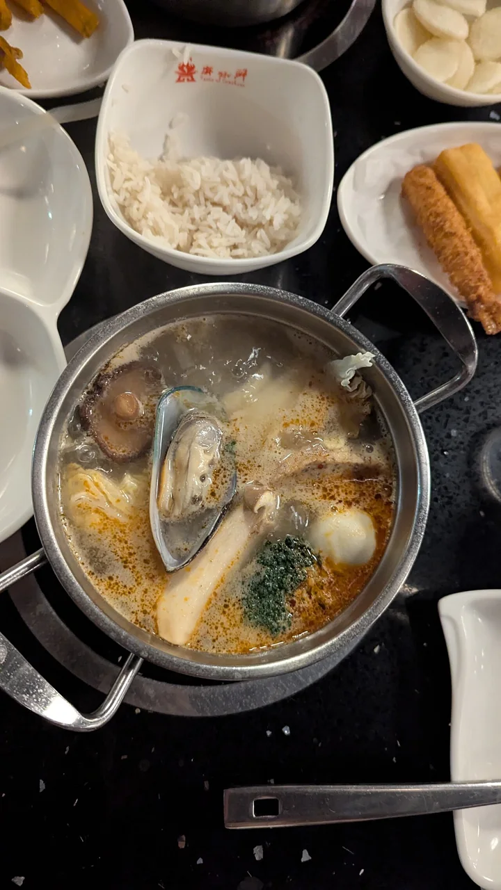 A metal pot of bubbling hotpot broth holds mussels, mushrooms, and vegetables.