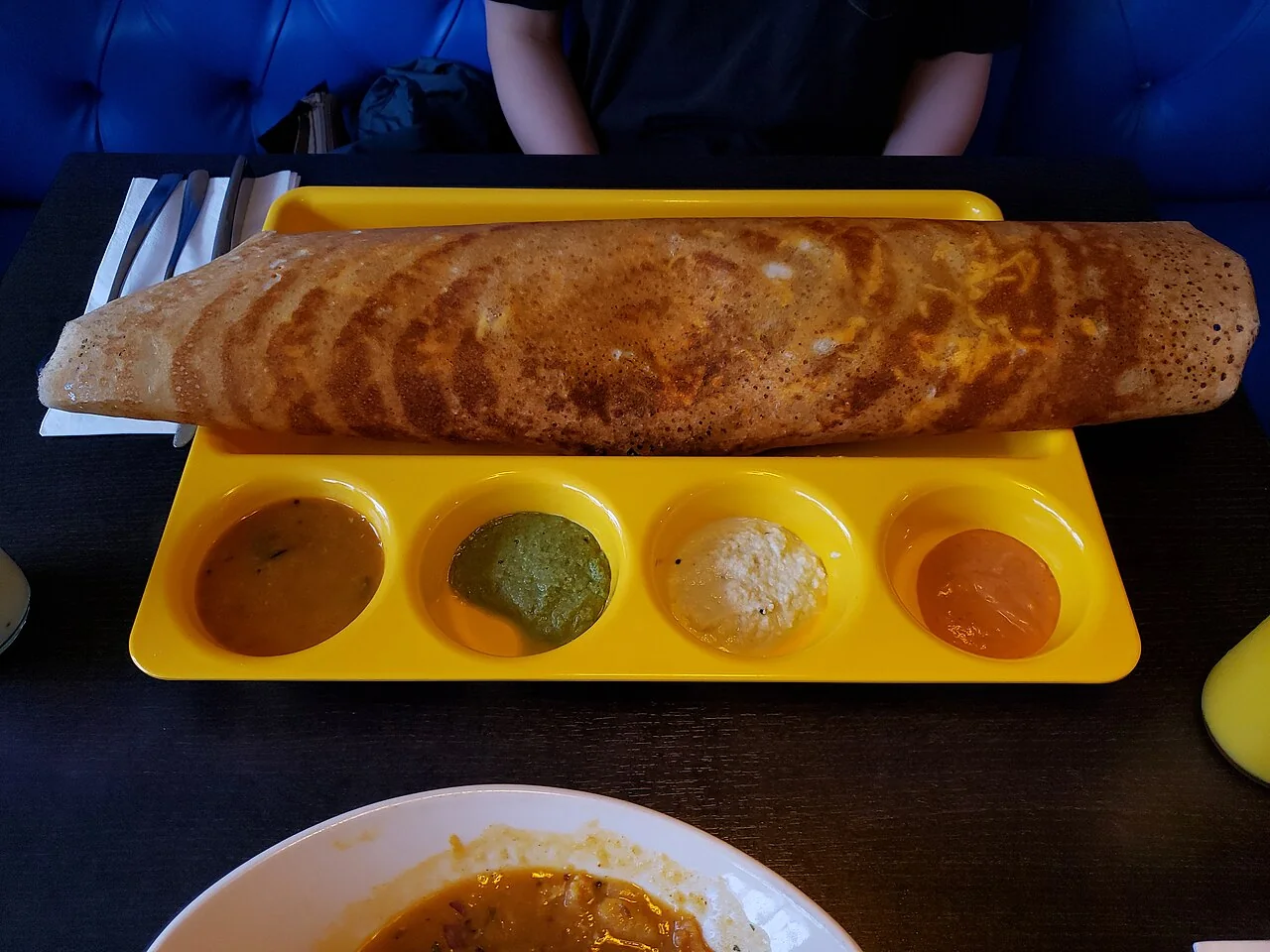 A long golden-brown thosai sits on a yellow tray with four small bowls of chutney.