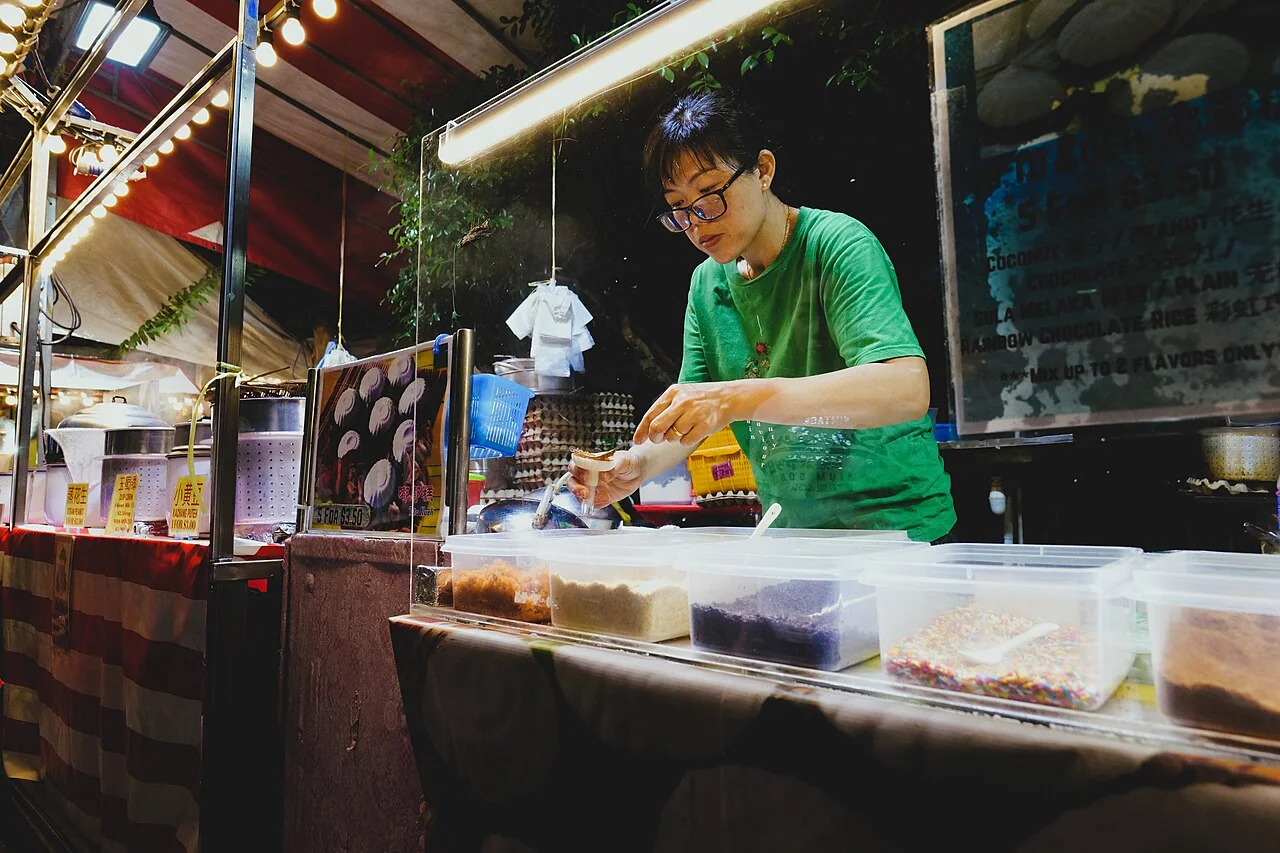 A vendor in a green shirt prepares tutu kueh at a brightly lit night market stall.
