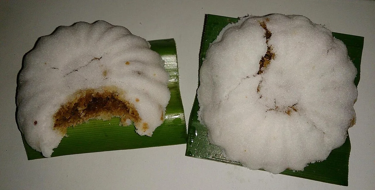 Two white tutu kueh with dark filling rest on pieces of green leaf.