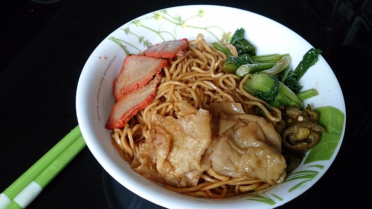 A white bowl holds thin noodles with red-edged pork slices, green leafy vegetables and flat dumplings, with green chopsticks nearby.