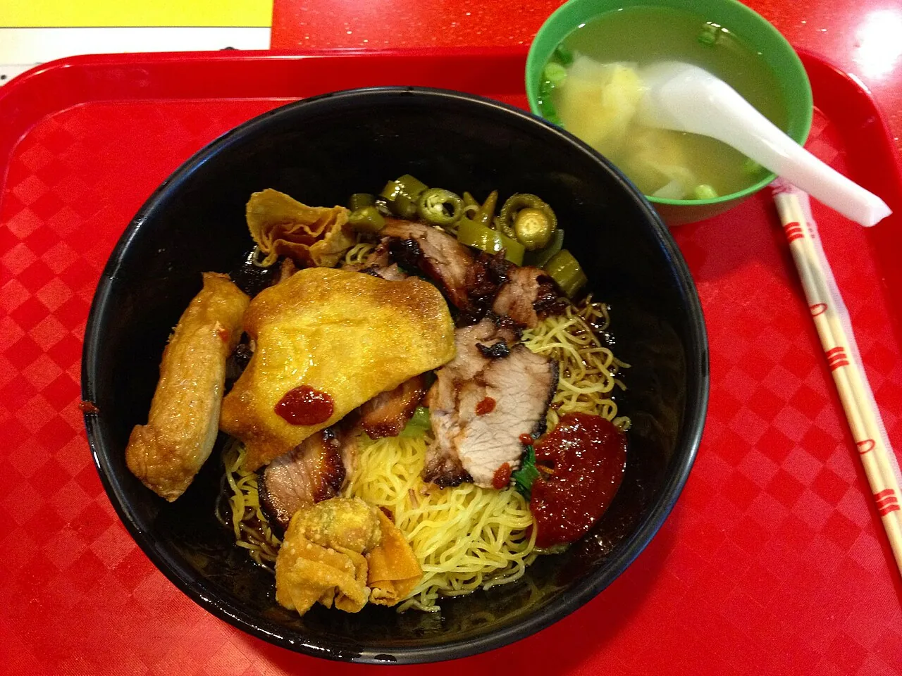 A black bowl of thin noodles topped with sliced pork, fried dumpling and green chillies, beside a small bowl of soup.