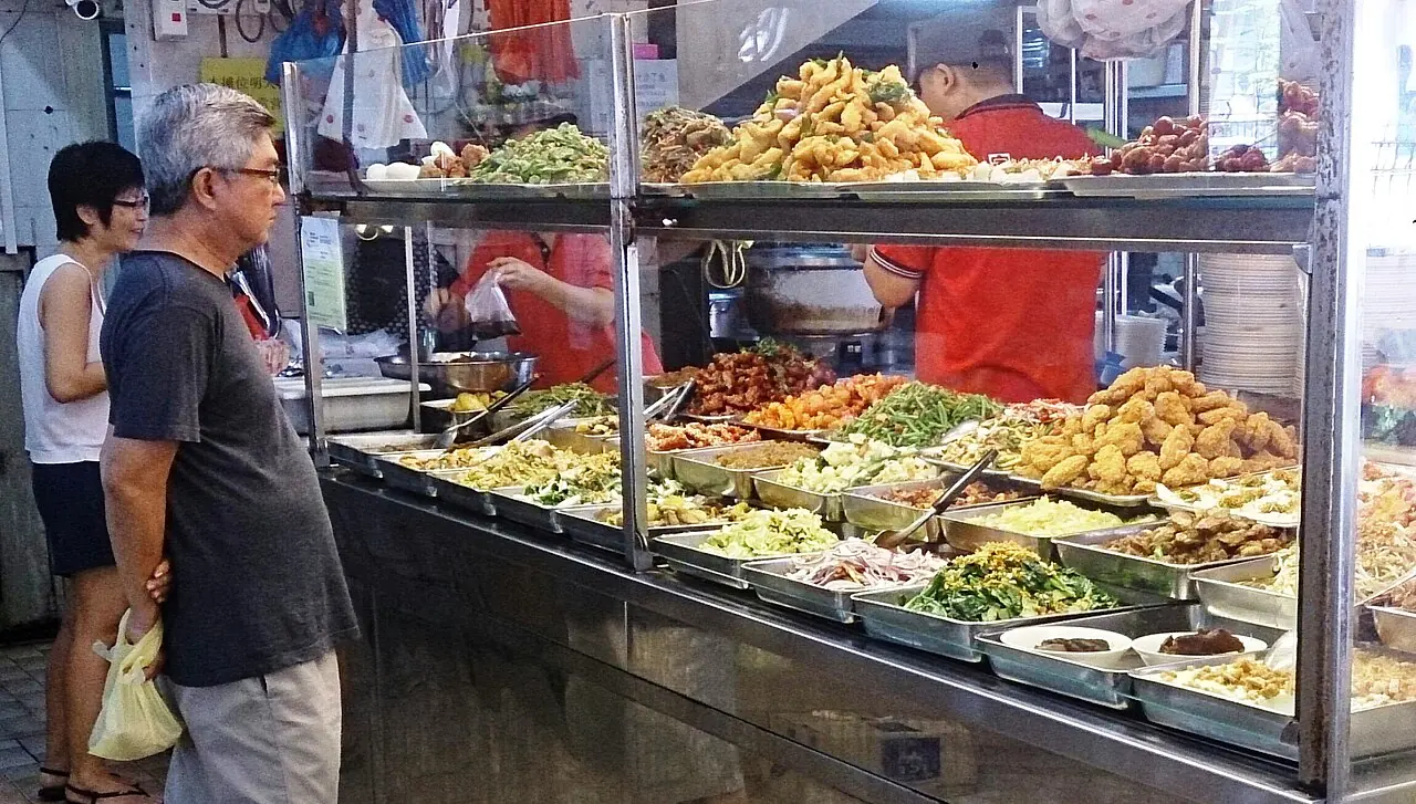 Customers queue beside trays of cooked dishes behind a rice-stall glass counter.