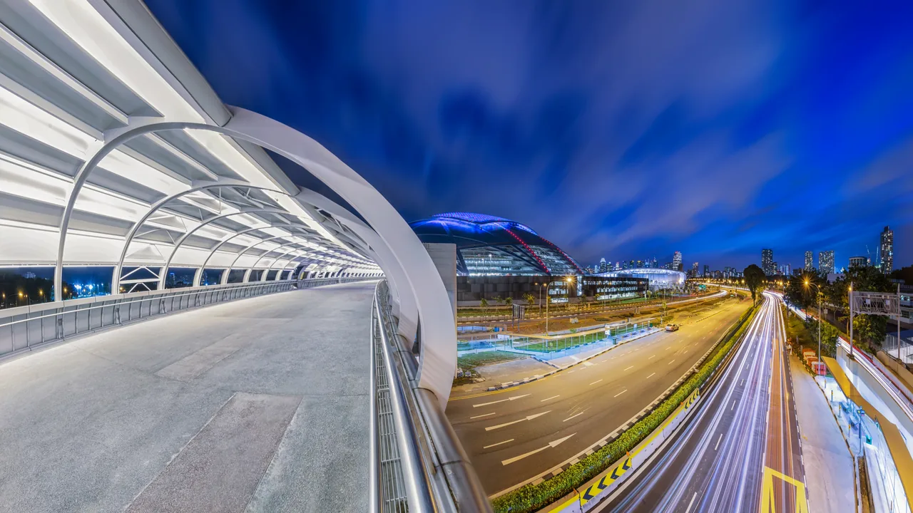 A curved pedestrian bridge and lit paths sweep through the sports hub.