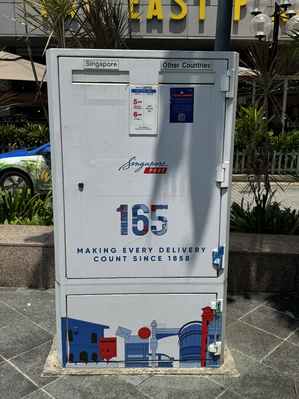 A white SingPost post box stands upright beside a pavement.