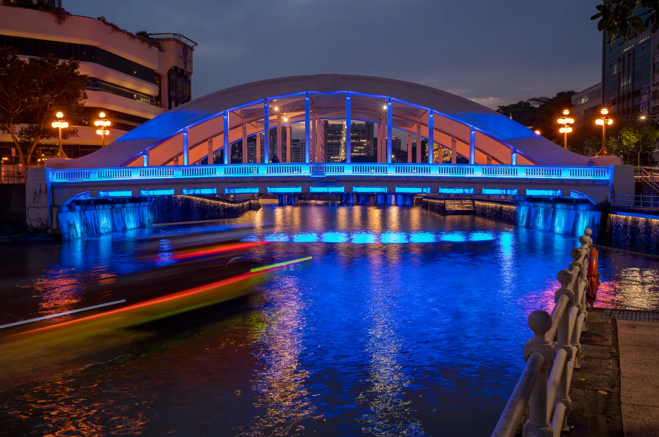 A blue-lit bridge spans the river while a boat passes below.