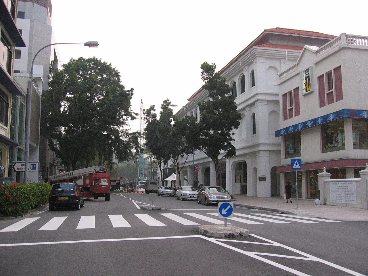 A street with a zebra crossing in the foreground, a blue pedestrian sign, and cars parked along the road.