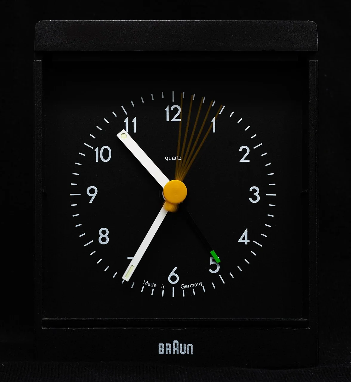 A black Braun quartz alarm clock with white numerals and a yellow centre cap is shown against a dark background.