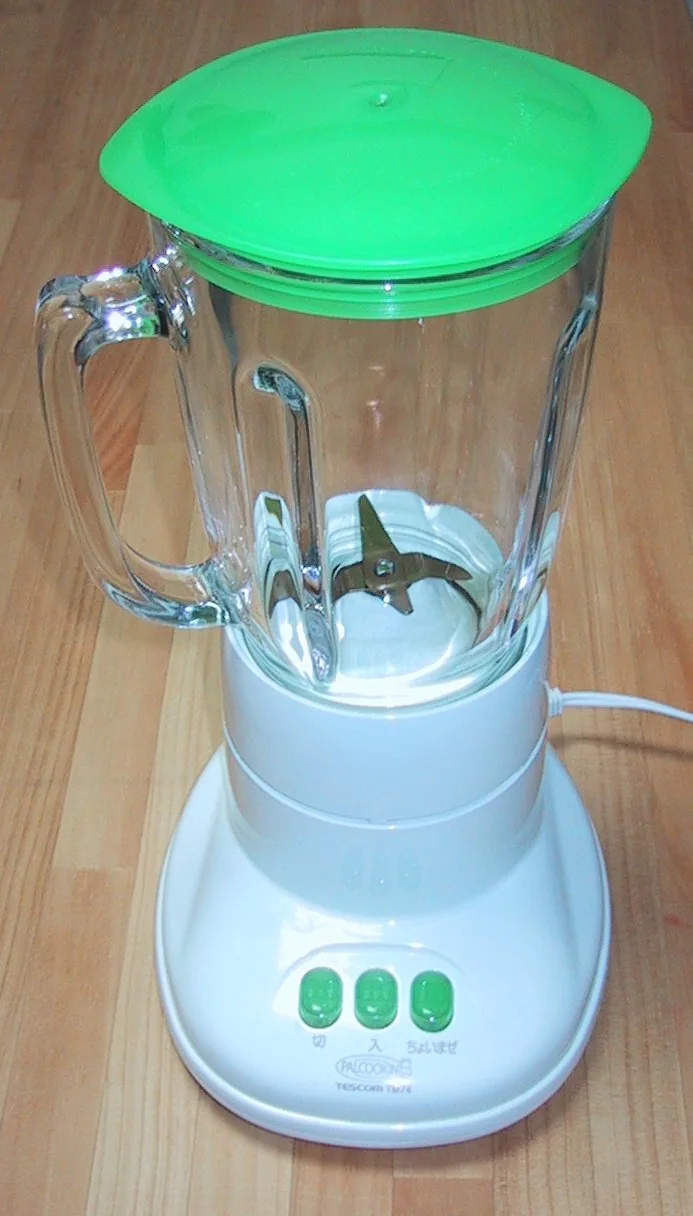 A white blender with a clear glass pitcher, green lid, and three green buttons sits on a wooden floor.