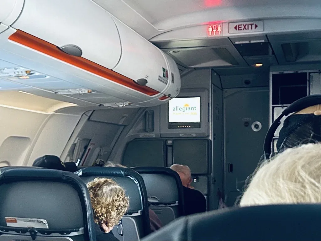 Passengers sit in the cabin of an Allegiant Air plane beneath overhead bins.