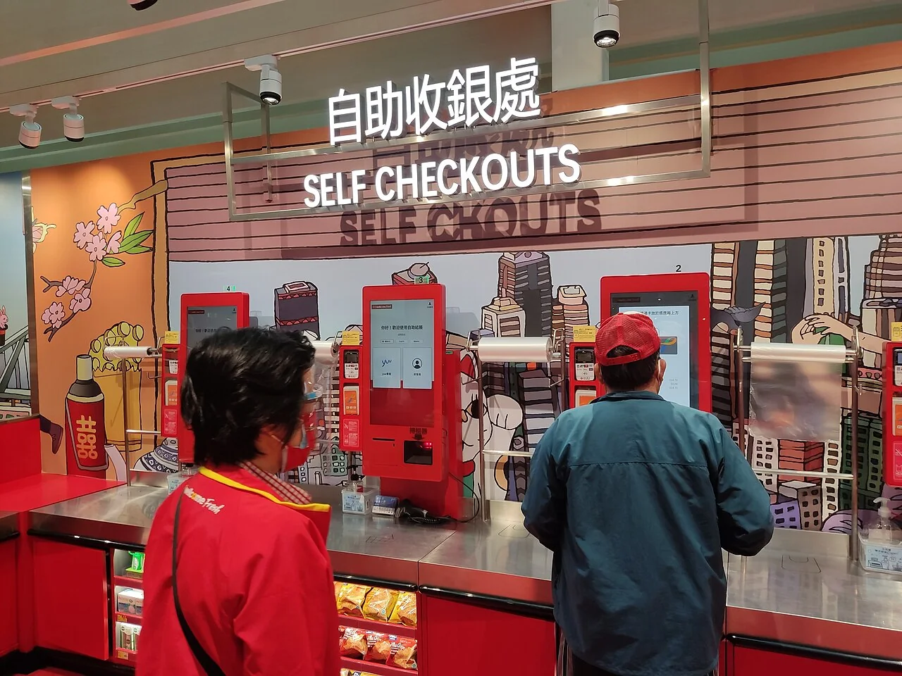 Two people stand at red self-checkout kiosks beneath a sign reading 'SELF CHECKOUTS'.
