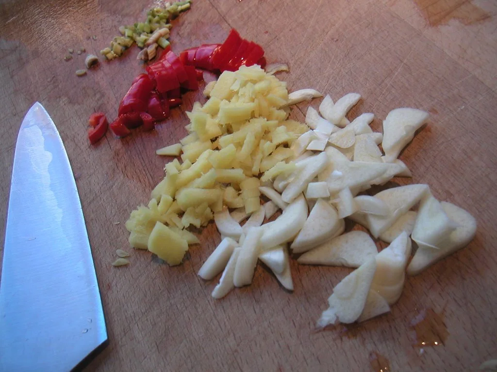 Chopped lemongrass, red chili, ginger and garlic are piled on a wooden board beside a knife.