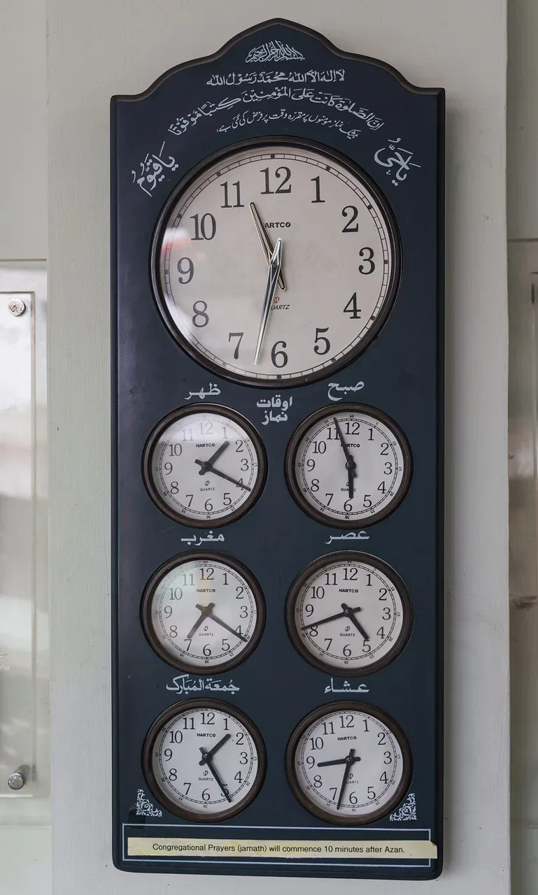 A multi-dial clock board showing prayer times hangs on a wall inside Jamae Mosque in Singapore.