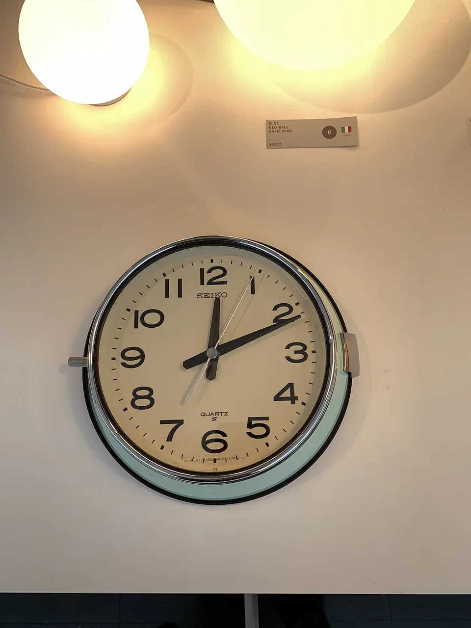 A Seiko quartz wall clock hangs on a white wall beneath two round ceiling lamps.